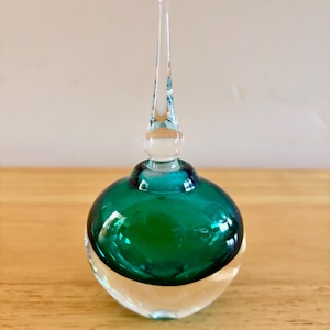 May include: A glass perfume bottle featuring a rounded base with a clear bottom section and a deep emerald green upper body. The bottle is topped with a tall, slender, clear glass stopper that tapers to a point, creating a sleek and smooth silhouette.