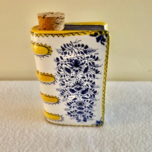 May include: A rectangular ceramic bottle with a cork stopper. The bottle is white with a blue floral design and yellow accents. The edges are outlined in yellow, and the cork is a light brown colour.