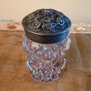 May include: A clear, faceted glass vase with a decorative metal lid. The lid is dark gray and features a floral design. The vase has a geometric pattern and is likely used for holding flowers or as a decorative piece.