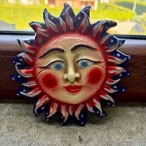 May include: A ceramic sun wall plaque featuring a central face with blue eyes, red cheeks, and red lips. The sun rays are painted in alternating shades of deep blue, bright red, and white, with small white dots decorating the blue sections.