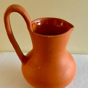 May include: A terracotta-colored ceramic pitcher with a rounded body, a wide opening, and a single tall, curved handle attached to the side. The exterior has a matte finish, while the interior shows a glossy, smooth glaze in a matching tone.