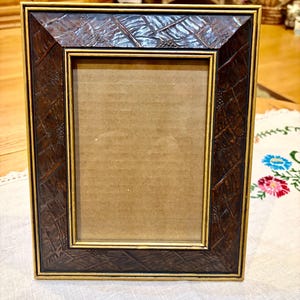 May include: A rectangular picture frame featuring a dark brown, textured border with a cross-hatched pattern and small raised details. The frame is accented with thin gold-toned trim along the inner and outer edges, surrounding a plain cardboard insert.