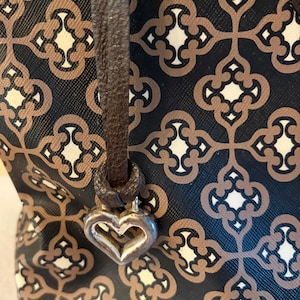 May include: A patterned handbag with a brown leather strap and a silver heart-shaped charm. The bag features a repeating geometric design in brown, cream, and black. The strap is attached to the bag with a metal ring.