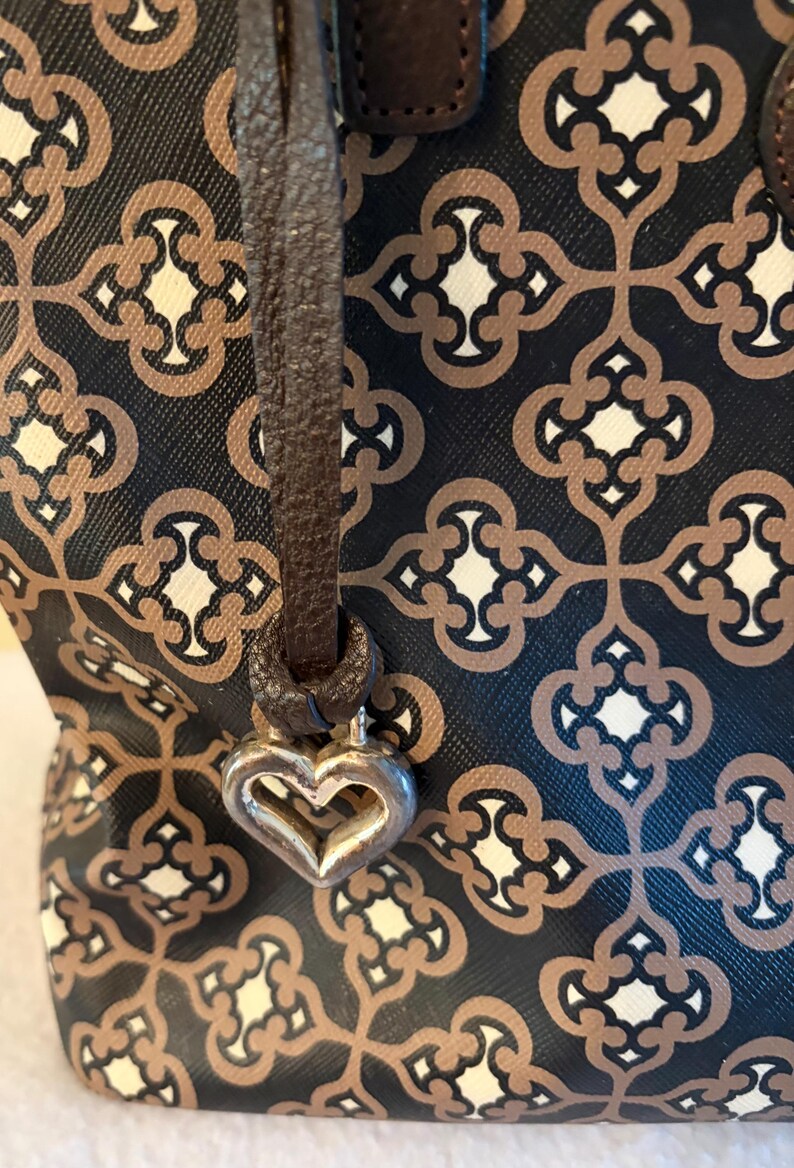 May include: A patterned handbag with a brown leather strap and a silver heart-shaped charm. The bag features a repeating geometric design in brown, cream, and black. The strap is attached to the bag with a metal ring.