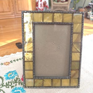 May include: A rectangular picture frame featuring a border of square, iridescent yellow-gold tiles. The frame is accented with a dark, textured metal trim along the inner and outer edges, surrounding a plain, light-colored rectangular insert.