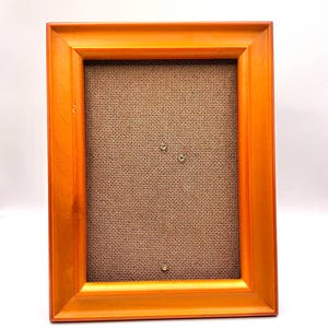 May include: A rectangular wooden picture frame with a warm orange finish. The frame has a simple, classic design with a textured, brown backing. The frame is designed to hold a photograph or artwork.