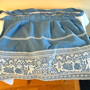 May include: A blue apron with a white folk art design along the bottom edge. The apron has a gathered waist and long ties. The white design features figures in traditional clothing and floral patterns. The apron is made of a textured fabric.