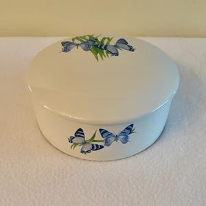 Puede incluir: Un recipiente redondo de cerámica blanca con tapa, que presenta un patrón decorativo de mariposas azules y briznas de hierba verde. El diseño está impreso tanto en la parte superior de la tapa como en el lateral de la base, con acabado brillante.