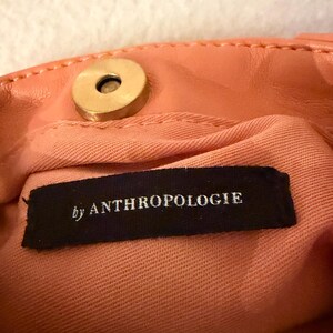 K&ouml;nnte beinhalten: Nahaufnahme des Innenraums einer pfirsichfarbenen Handtasche mit einem goldfarbenen Magnetverschluss. Das Innenfutter ist ein hellerer pfirsichfarbener Stoff. Ein schwarzes Etikett mit dem Text "by ANTHROPOLOGIE" ist sichtbar.