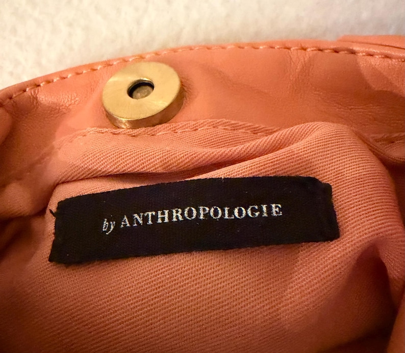 K&ouml;nnte beinhalten: Nahaufnahme des Innenraums einer pfirsichfarbenen Handtasche mit einem goldfarbenen Magnetverschluss. Das Innenfutter ist ein hellerer pfirsichfarbener Stoff. Ein schwarzes Etikett mit dem Text "by ANTHROPOLOGIE" ist sichtbar.