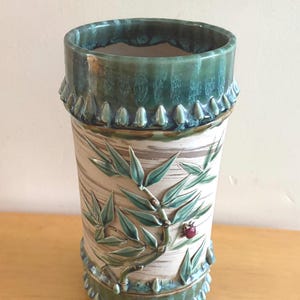 Könnte beinhalten: Eine zylindrische Keramikvase mit cremefarbenem Körper und einem Reliefdesign aus Bambusstängeln und Blättern. Der obere und untere Rand sind mit einer glänzenden dunkelgrünen Glasur und einem dekorativen Wellenrand versehen. Ein kleiner roter Marienkäfer ist am Bambusstängel befestigt.