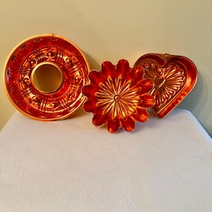May include: Three copper-colored vintage baking molds. A round, fluted bundt pan, a flower-shaped mold, and a heart-shaped mold. The molds have intricate embossed designs and are displayed on a white surface.