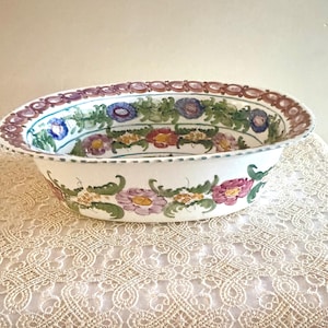 May include: Oval ceramic serving bowl with a hand-painted floral design. The bowl features a white base with a border of purple, blue, and red flowers and green leaves. The bowl is placed on a cream-colored lace tablecloth.