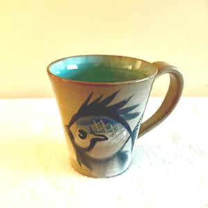 May include: A ceramic mug featuring a fish design. The mug has a light brown exterior with a blue and white fish illustration. The interior is teal green. The handle is light brown. A unique piece for tea or coffee.