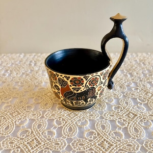 Puede incluir: Taza de cerámica pequeña con asa negra e interior negro. El exterior presenta un fondo color crema con un patrón repetido de diseños florales y animales rojos y negros. El asa arqueada está unida a la taza.