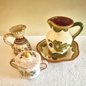 May include: A set of three ceramic pieces: a small pitcher, a covered sugar bowl, and a pitcher with a matching plate. The pieces are cream-colored with brown, green, and yellow painted floral designs. The pitcher has a green handle.