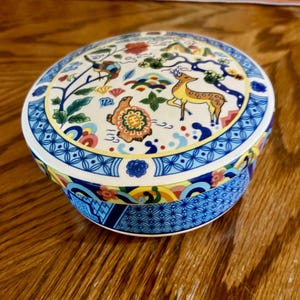 May include: A round ceramic lidded box featuring a colorful painted design of a deer, a turtle, and stylized trees. The lid and base are decorated with intricate blue and white geometric patterns, along with accents of red, green, and yellow.