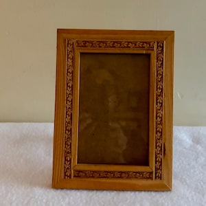 Może przedstawiać: Prostokątna drewniana ramka na zdjęcia z jasnobrązowym wykończeniem. Ramka ma dekoracyjny kwiatowy wzór w ciemniejszym czerwonobrązowym kolorze wzdłuż krawędzi. Ramka jest pusta, z ciemnym wnętrzem.