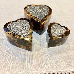 Puede incluir: Tres cajas en forma de corazón con un patrón de carey marrón y negro. Cada tapa presenta una placa de metal plateado incrustada con un intrincado diseño floral y de volutas en relieve. Las cajas son de diferentes tamaños.