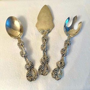 May include: A trio of silver-coloured serving utensils: a spoon, a pie server, and a fork. Each handle features an elaborate grape vine design, with leaves and bunches of grapes. The spoon has a rounded bowl, the server has a triangular blade, and the fork has two prongs.