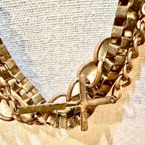 May include: A multi-layered necklace featuring a thick, metallic box chain and a thinner, rounded link chain. A small, simple metallic cross pendant is attached to the lower section of the chain, creating a layered look with a weathered, antique finish.