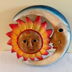 May include: A carved wooden wall plaque featuring a central sun face with yellow and red rays, partially encircled by a blue and white crescent moon face. The sun has a natural wood tone, while the moon has a smooth, curved profile.