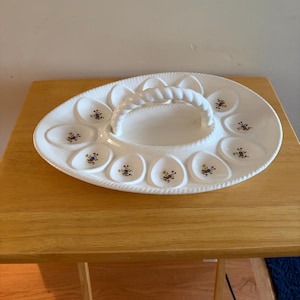 May include: A white ceramic egg serving platter with an oval shape and a central twisted handle. The platter features twelve individual egg-shaped indentations, each decorated with a small, colorful floral pattern of blue, yellow, and green.