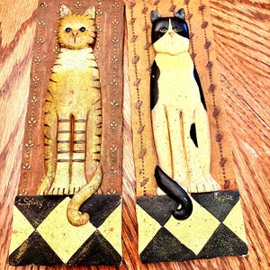 May include: Two rectangular wall plaques featuring sculpted cats. The left plaque shows a yellow tabby cat on a brown background with vertical leaf patterns. The right plaque shows a black and white cat. Both have black and yellow diamond bases.