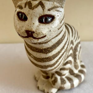 May include: A ceramic cat figurine featuring a cream-colored base with dark brown striped patterns. The cat has pointed ears, dark brown eyes, and a painted nose and mouth. The surface has a speckled texture, and the cat is depicted in a seated position.