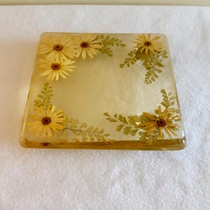 Può includere: Un sottobicchiere quadrato in resina trasparente con fiori di margherita gialli pressati e delicate foglie di felce verde incastonate lungo il bordo, creando un motivo botanico all'interno del materiale liscio, trasparente e lucido.