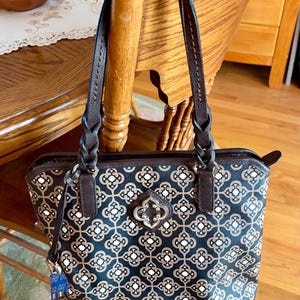 May include: A patterned tote bag with brown leather handles and accents. The bag features a repeating floral design in black, tan, and white. The handles are braided and attached to the bag with metal hardware.