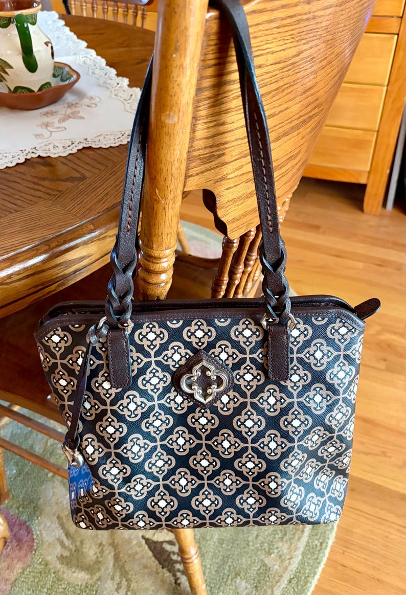 May include: A patterned tote bag with brown leather handles and accents. The bag features a repeating floral design in black, tan, and white. The handles are braided and attached to the bag with metal hardware.