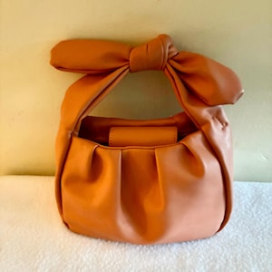 K&ouml;nnte beinhalten: Eine kleine, orangefarbene Handtasche mit einem geknoteten Henkel. Die Tasche hat eine runde Form mit Falten am Boden und einen kleinen, rechteckigen Klappenverschluss. Der Henkel ist ein gro&szlig;er, dekorativer Knoten. Die Tasche ist aus glattem, leder&auml;hnlichem Material.