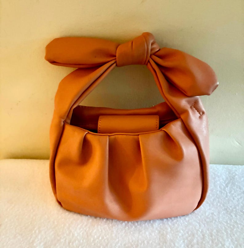 K&ouml;nnte beinhalten: Eine kleine, orangefarbene Handtasche mit einem geknoteten Henkel. Die Tasche hat eine runde Form mit Falten am Boden und einen kleinen, rechteckigen Klappenverschluss. Der Henkel ist ein gro&szlig;er, dekorativer Knoten. Die Tasche ist aus glattem, leder&auml;hnlichem Material.