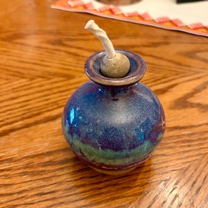 May include: A small, spherical ceramic oil lamp with a glossy glaze in shades of deep blue, purple, and green. The lamp features a narrow neck with a flared rim and a central stone-colored stopper holding a thick, off-white cotton wick.