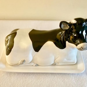 Puede incluir: Una mantequillera de cerámica con forma de vaca reclinada con un acabado brillante de manchas blancas y negras. La figura de la vaca sirve como tapa, descansando sobre una bandeja base rectangular blanca con bordes elevados y superficie lisa.