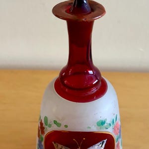 May include: A glass perfume bottle with a deep red stopper and neck. The frosted white body features a central oval medallion with a painted butterfly, surrounded by hand-painted floral accents in green, red, and blue, with gold trim.