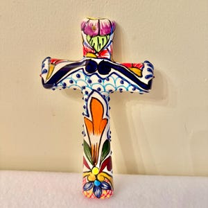 Może przedstawiać: Ręcznie malowany ceramiczny krzyż z żywym kwiatowym wzorem. Krzyż ma białą podstawę z niebieskimi, pomarańczowymi, czerwonymi, żółtymi i fioletowymi akcentami. Ozdobny krzyż ma około 15 cm wysokości.