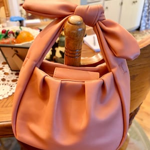K&ouml;nnte beinhalten: Eine blush-rosa Handtasche mit einem geknoteten Griff. Die Tasche hat ein gerafftes Design auf der Vorderseite und einen Klappenverschluss. Der Griff ist zu einer gro&szlig;en Schleife gebunden. Die Tasche liegt auf einem Holzstuhl.