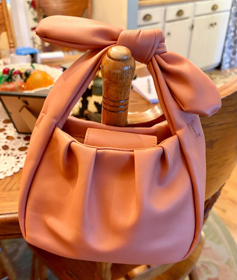 K&ouml;nnte beinhalten: Eine blush-rosa Handtasche mit einem geknoteten Griff. Die Tasche hat ein gerafftes Design auf der Vorderseite und einen Klappenverschluss. Der Griff ist zu einer gro&szlig;en Schleife gebunden. Die Tasche liegt auf einem Holzstuhl.
