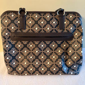 May include: A rectangular tote bag with a repeating geometric pattern in black, brown, and white. The bag has two brown handles attached with silver rings, a brown zipper pocket, and a brown trim.