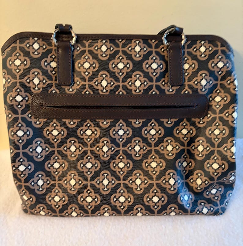 May include: A rectangular tote bag with a repeating geometric pattern in black, brown, and white. The bag has two brown handles attached with silver rings, a brown zipper pocket, and a brown trim.