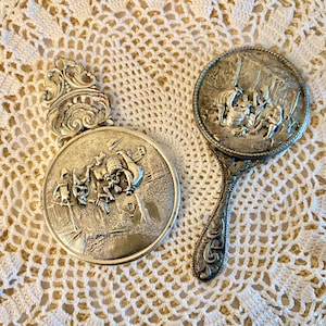 May include: Two antique silver-toned compact mirrors with detailed relief scenes. One is round with a handle, the other is a round compact with an ornate top. Both are on a white lace doily.