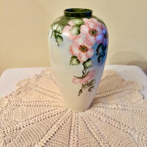 May include: A white ceramic vase with a painted floral design. The vase features pink flowers with green leaves and a dark green rim. The vase is set on a cream-colored, crocheted doily.
