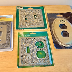 May include: Four packaged wall plates including a rectangular single toggle switch plate, a square double toggle switch plate, a square combination toggle and outlet plate, and an oval duplex outlet plate, all featuring metallic finishes.