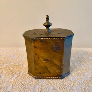 May include: An antique brass-toned metal container with an octagonal shape and a lid. The container has a decorative rope-like trim around the top and bottom. The lid is topped with a decorative finial. The container has a vintage, aged appearance.