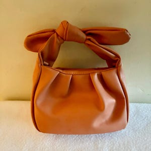 K&ouml;nnte beinhalten: Eine orangefarbene Handtasche mit einem geknoteten Griff. Die Tasche hat ein gerafftes Design auf der Vorderseite und einen bogenf&ouml;rmigen Griff. Die Tasche ist aus einem glatten Material und hat eine einfarbige orange Farbe.