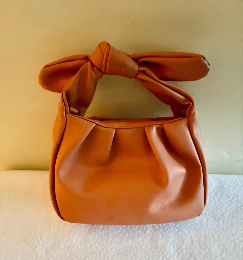 K&ouml;nnte beinhalten: Eine orangefarbene Handtasche mit einem geknoteten Griff. Die Tasche hat ein gerafftes Design auf der Vorderseite und einen bogenf&ouml;rmigen Griff. Die Tasche ist aus einem glatten Material und hat eine einfarbige orange Farbe.