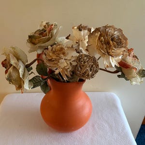 May include: A matte orange ceramic pitcher filled with a decorative arrangement of dried, off-white and brown-toned artificial flowers, green fabric leaves, and a small, spherical woven rattan orb nestled among the stems.