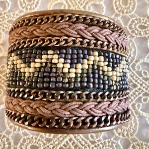 May include: A wide cuff bracelet with a mix of textures and colors. The bracelet features braided sections, metal chain links, and a beaded center with cream and dark blue beads. The overall color palette includes shades of brown, gold, and blue.